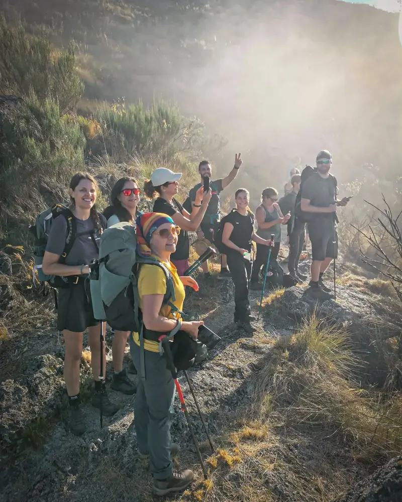Grupo de caminhantes da 100 Montanhas com mochilas e bastões num trilho de montanha, ao sol da manhã